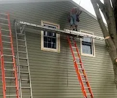 A worker stands on a makeshift platform created by resting a ladder horizontally across two extension ladders.