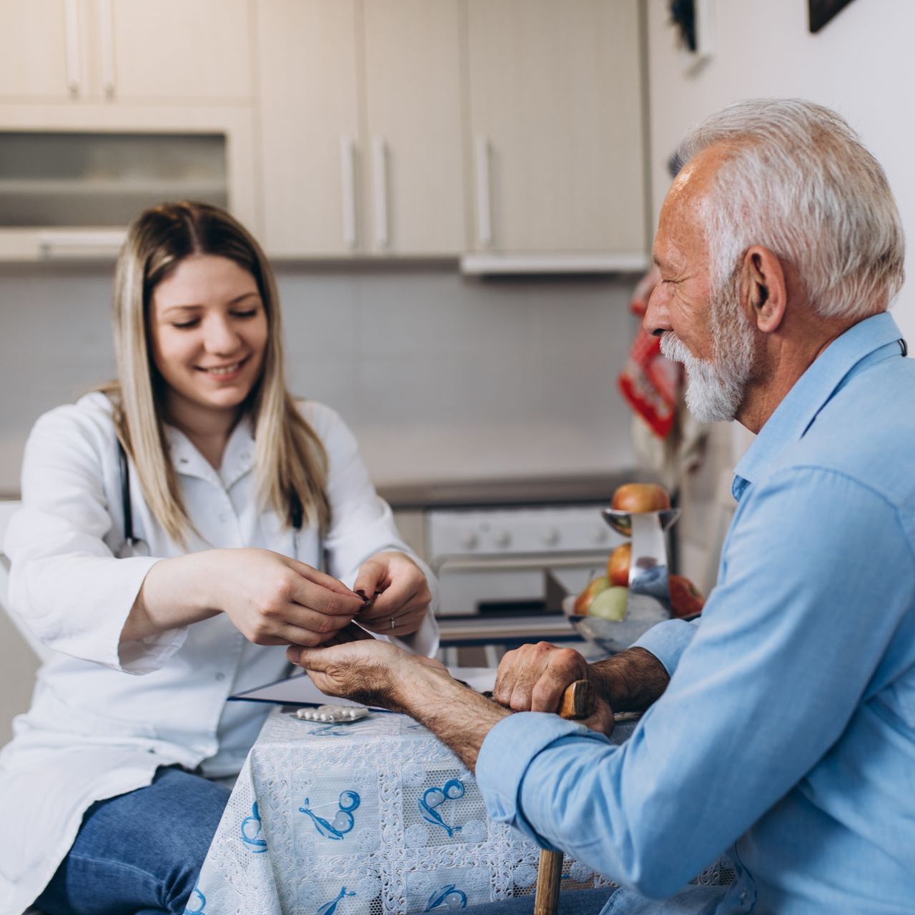 A female doctor checking an older man's wrist in a kitchen. Both smiling; doctor in white coat.
