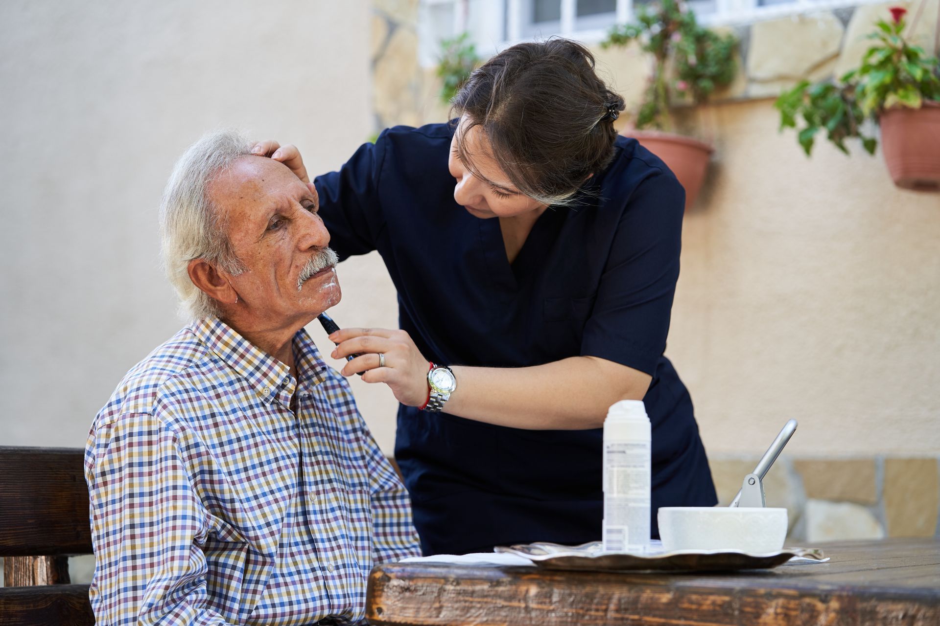 Woman shaving an elderly man's face outdoors at a table. The man is seated, and the woman stands next to him.