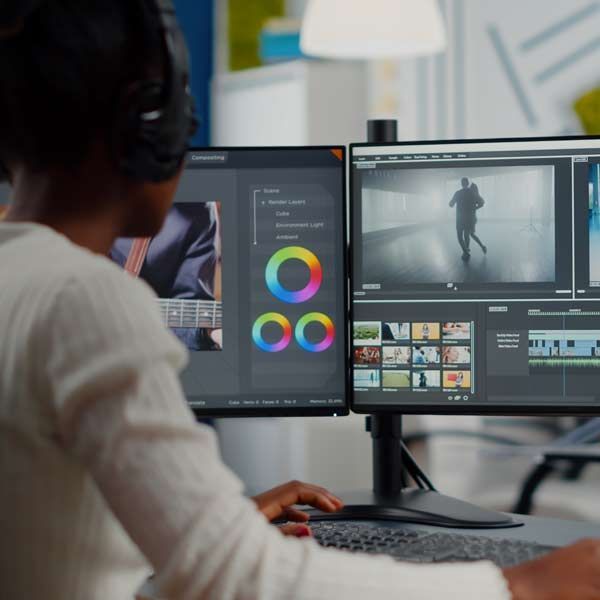 A video marketer wearing headphones edits a video on dual computer monitors.