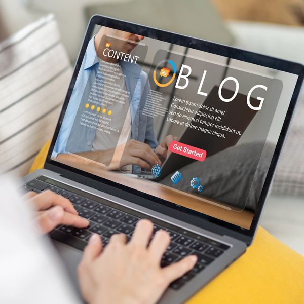 A blogger typing on a laptop, viewing a blog website on the screen, inside a room.