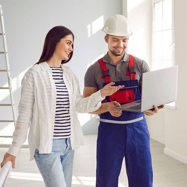 A woman and a general contractor are looking at a laptop in an empty room. He points; she smiles, holding blueprints.