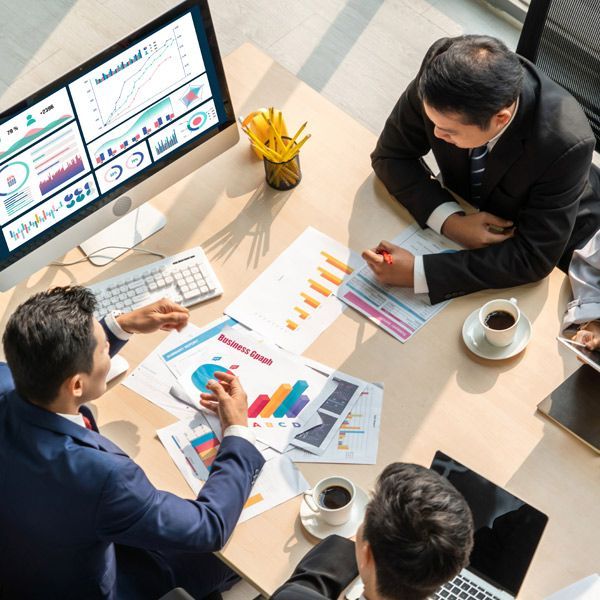 Business meeting around a table, analyzing marketing reports and graphs displayed on a monitor and on paper.