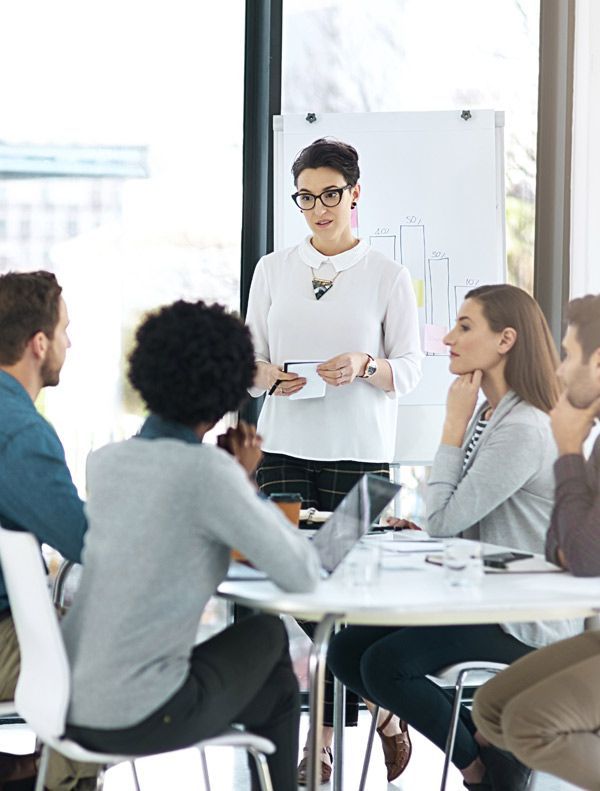 A woman leading an SEO meeting at SEO Agency Solution Web Designs, gesturing towards a whiteboard, with colleagues at a table.