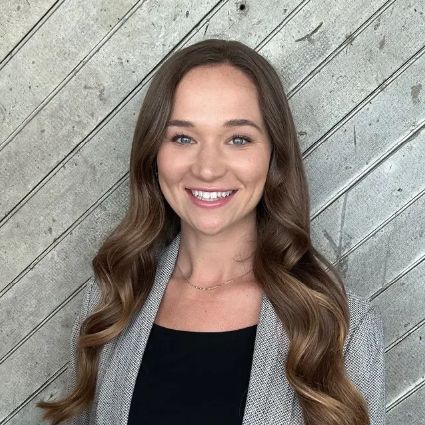 Smiling woman with long wavy hair in a gray cardigan against a diagonal wood wall