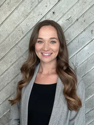 Smiling woman in a gray sweater standing against a diagonally paneled wood wall.