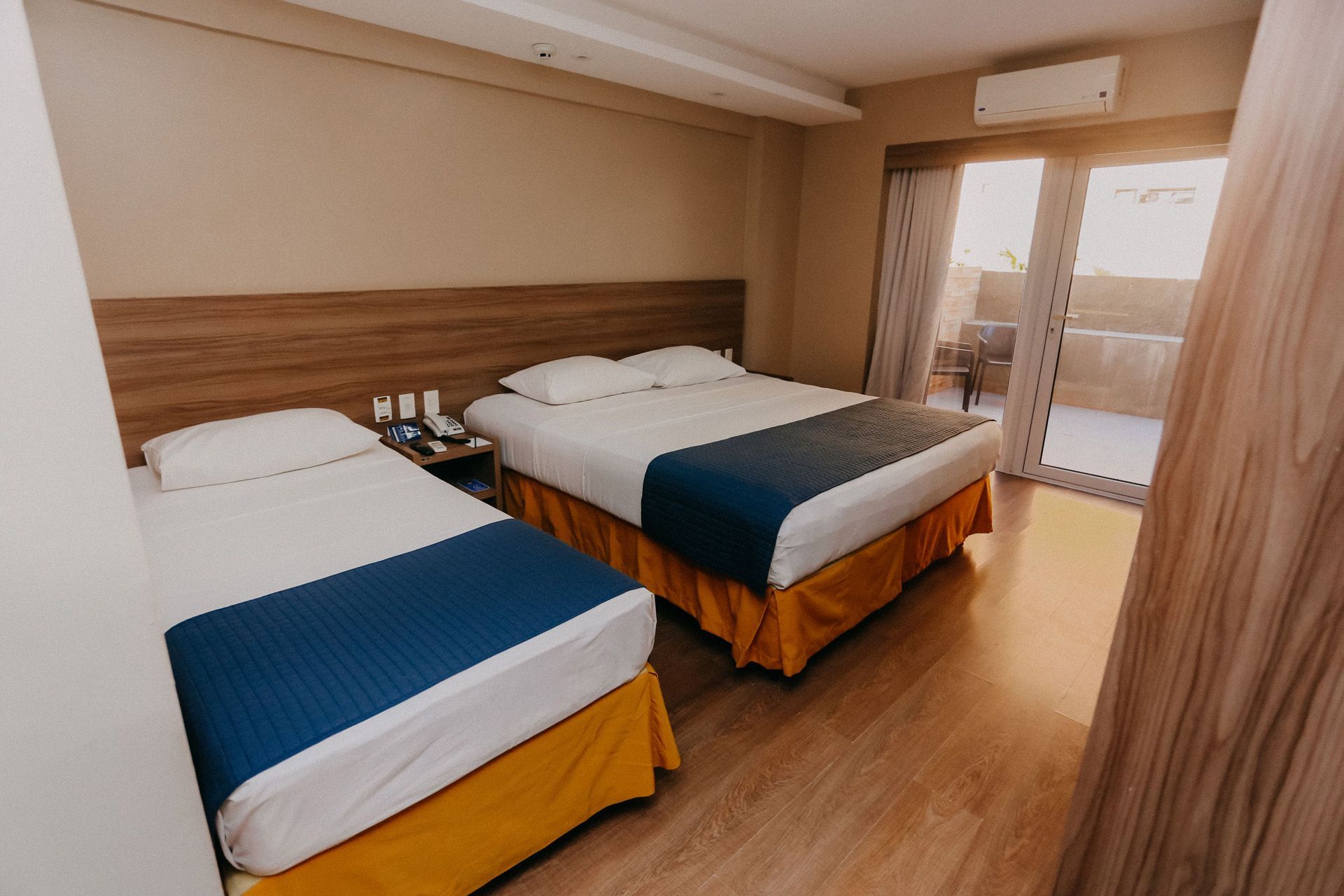 Quarto de hotel com duas camas, uma de casal e uma de solteiro, e uma varanda.