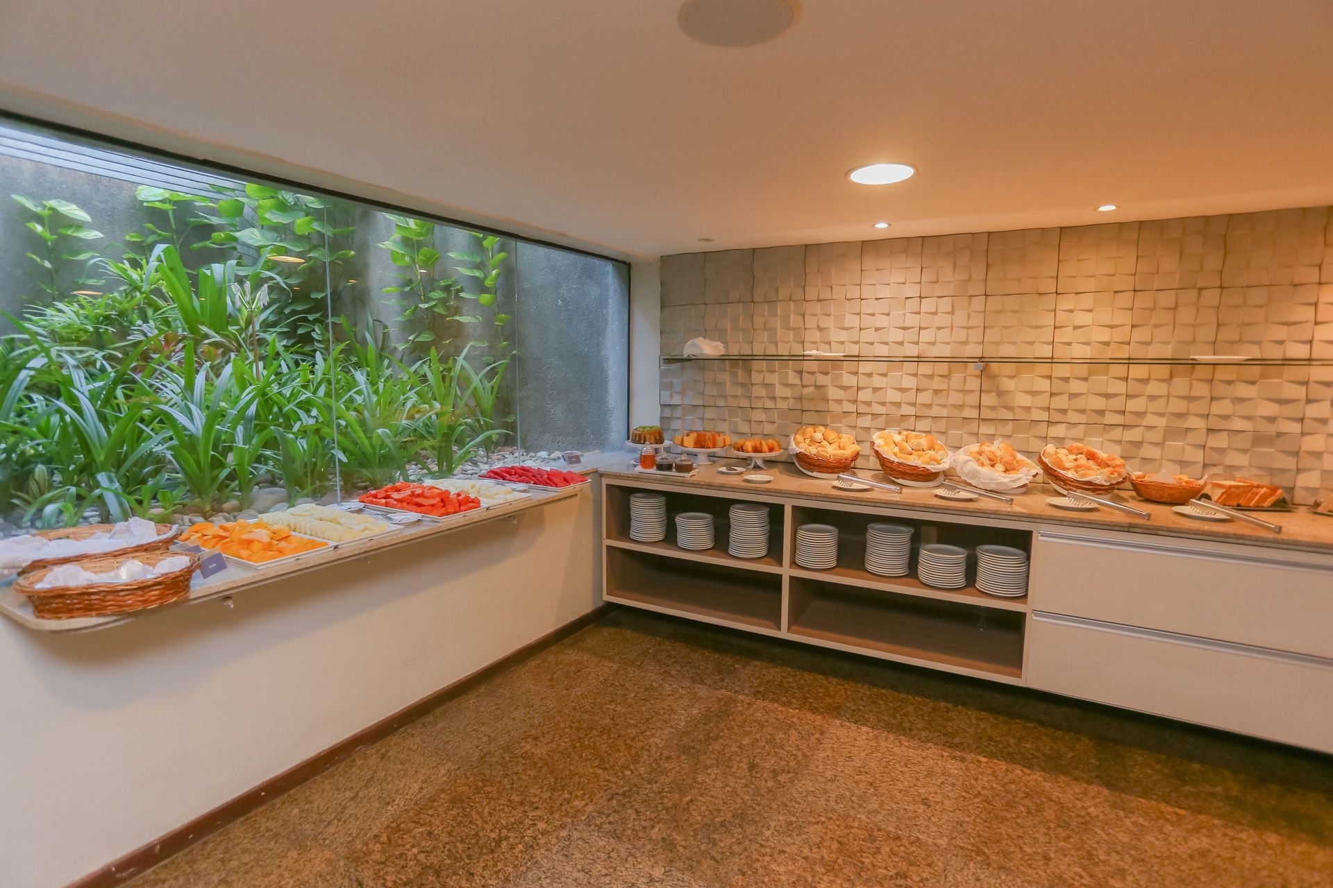 Buffet de café da manhã com diversos itens alimentares: pães, bolos, frutas e pratos quentes, em um refeitório.