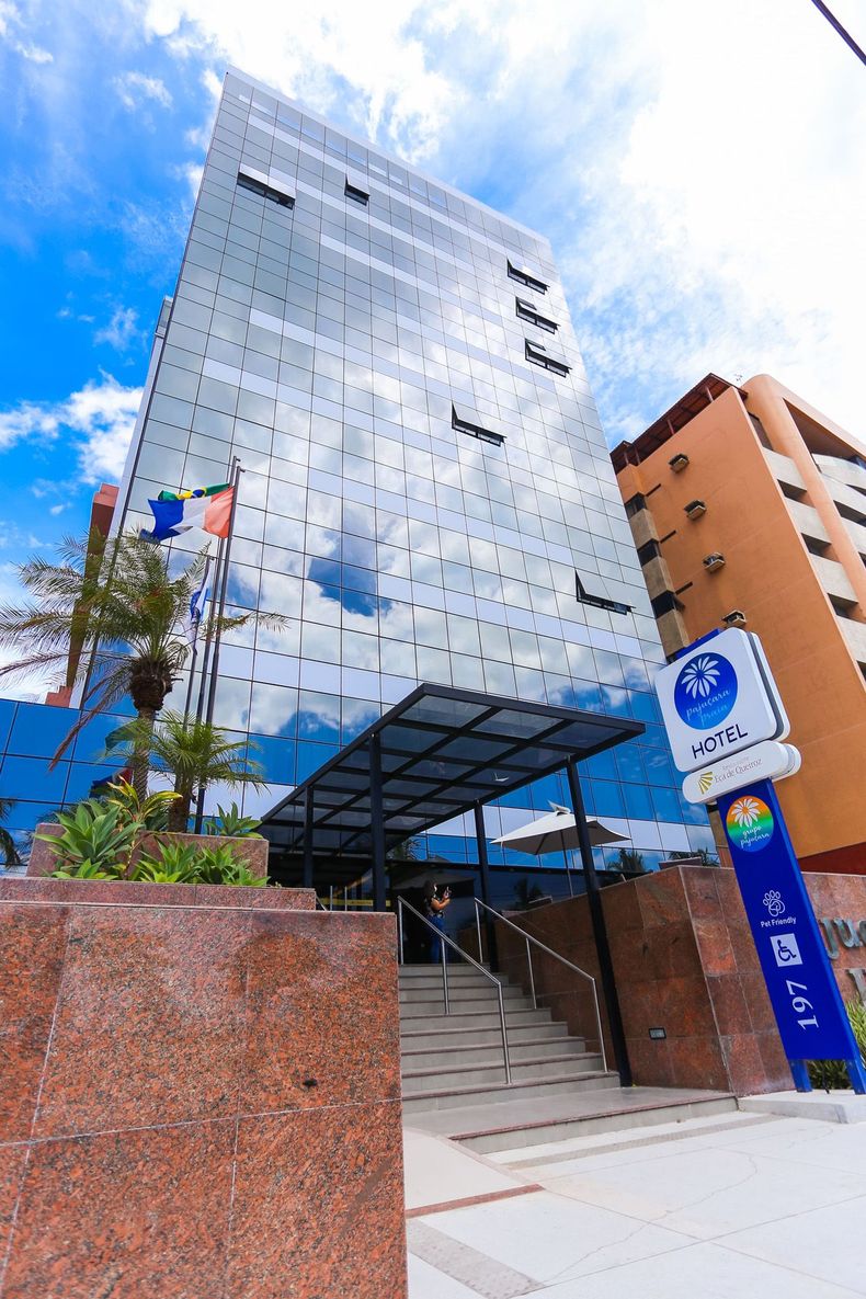 Prédio alto e moderno com fachada de vidro, refletindo o céu. Sinalização e escadaria de acesso visíveis.