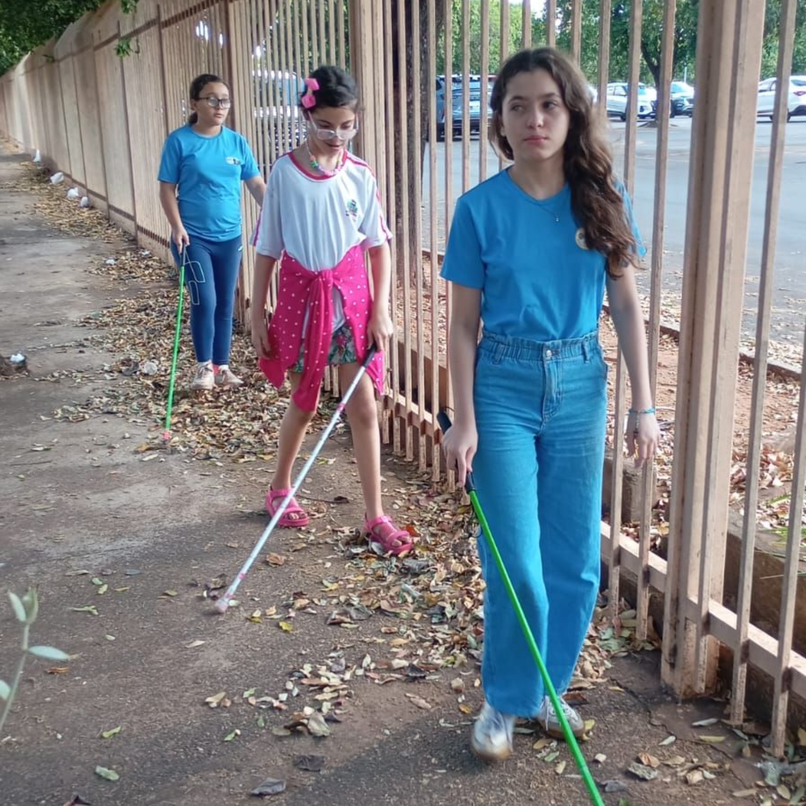 Uma criança caminhando em área externa segurando uma bengala longa, acompanhado por uma senhora que o segue logo atrás também segurando uma bengala longa.
