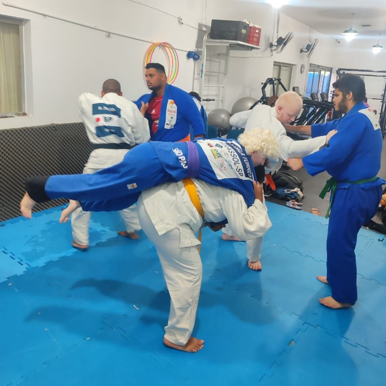 Grupo de seis pesoas de pé uma do lado da outra no tatame, vestindo kimono e máscaras faciais.