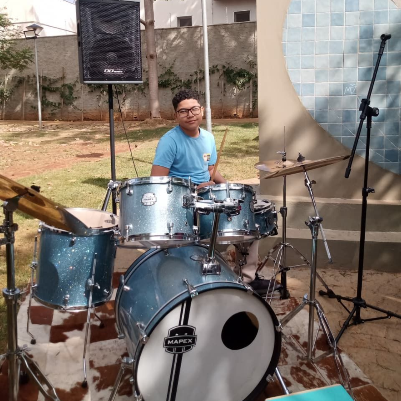 Menino usando óculos escuras e máscara facial tocando bateria com violões pendurados na parede ao fundo.