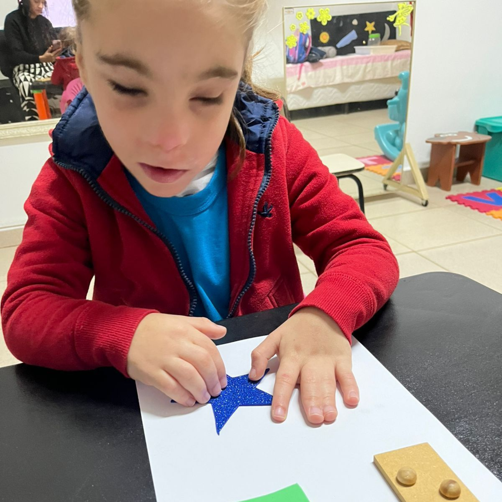 Menino sentato na carteira acompanhado da professora pedagógica que o auxilia na atividade de colorir um desenho.