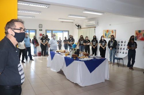 Um homem usando uma máscara está de pé em frente a uma mesa com pessoas ao redor dela.