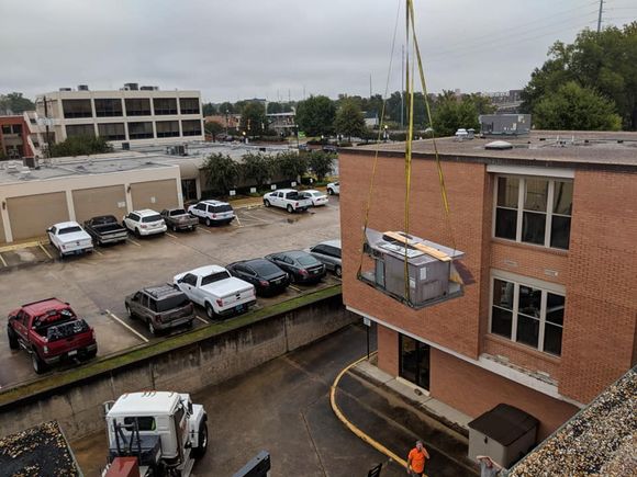 A crane lifting an HVAC unit onto a brick building with a parking lot and cars in view.