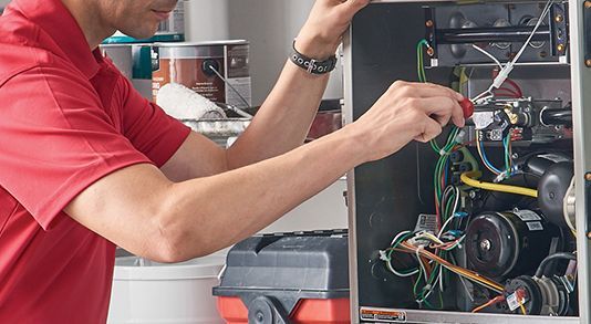 A man in a red shirt repairs a furnace, using a screwdriver.