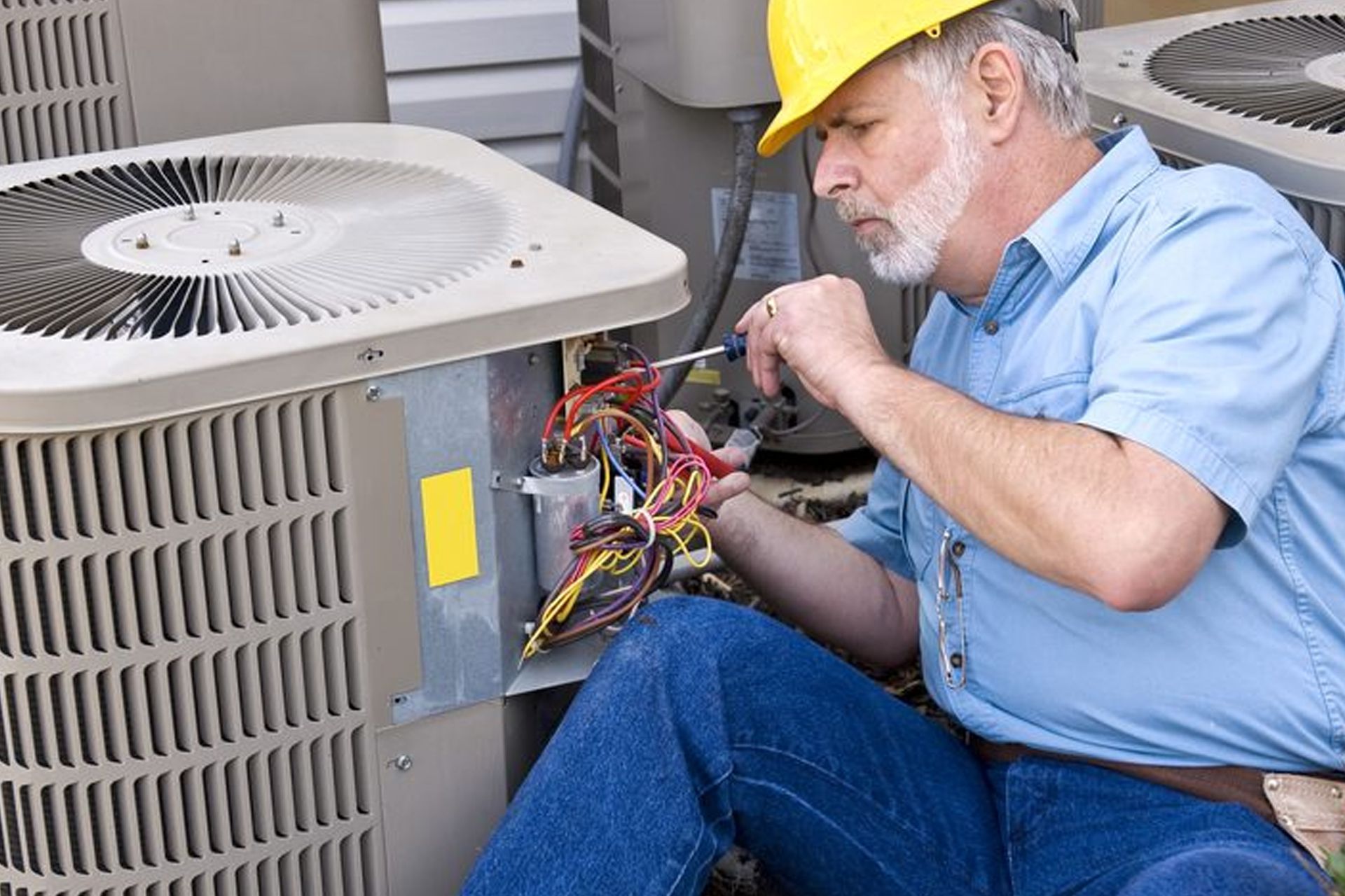 HVAC technician in yellow hard hat, working on outdoor air conditioning unit with screwdriver.