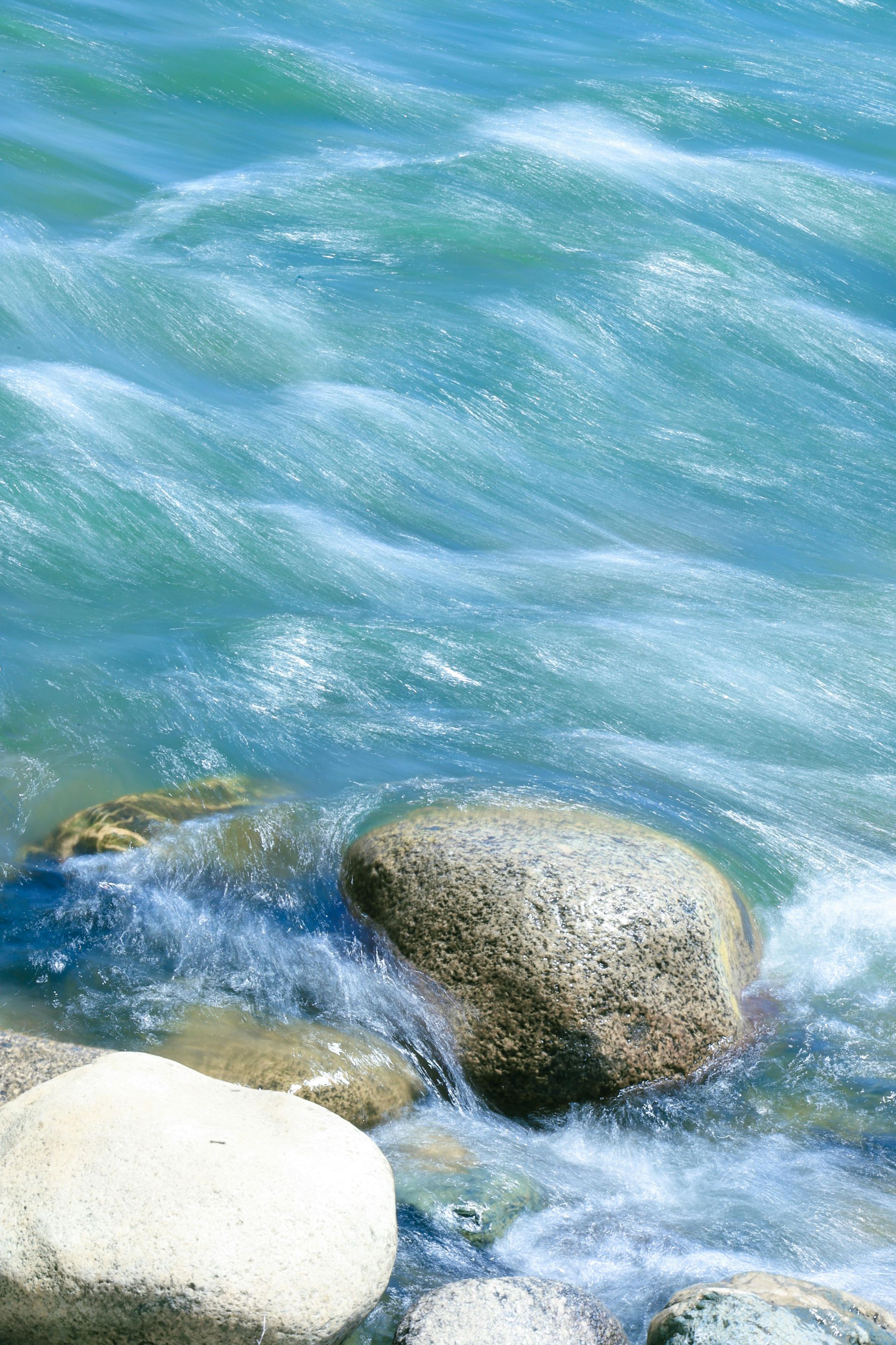 Fast-moving turquoise water flows over and around several smooth, rounded stones along a shoreline.
