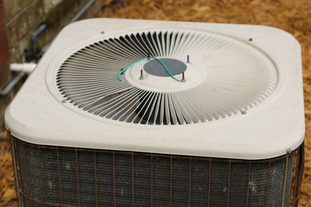 Outdoor air conditioning unit with fan, near a brick wall and a tree.