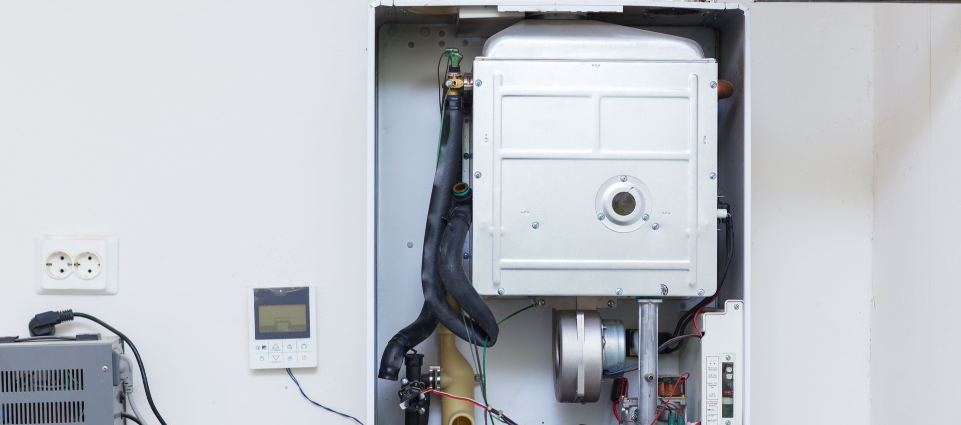 Open boiler with visible components, mounted on a white wall with an electrical outlet and thermostat.