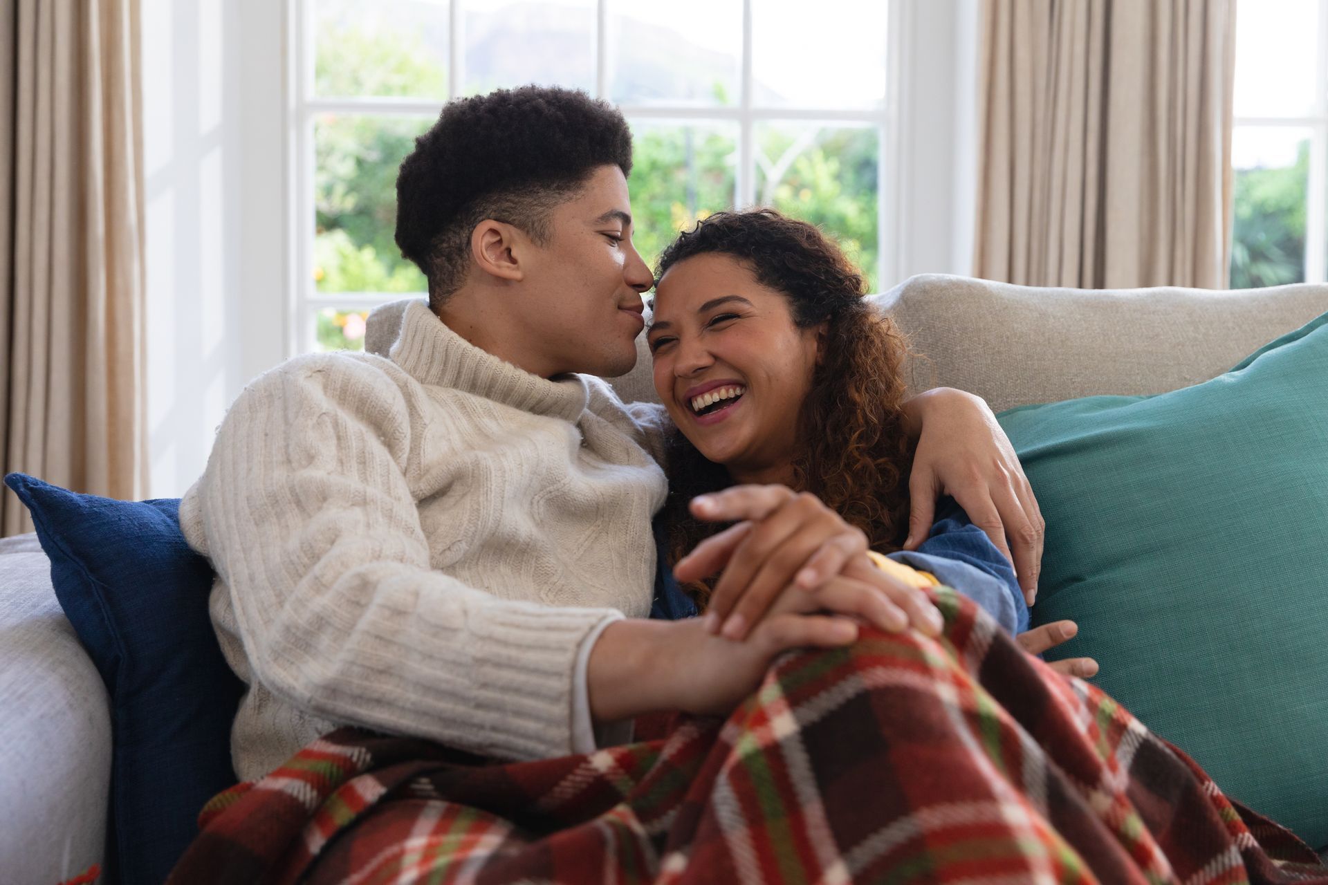 A couple laughs together on a couch, covered with a blanket. Man whispers in woman's ear.