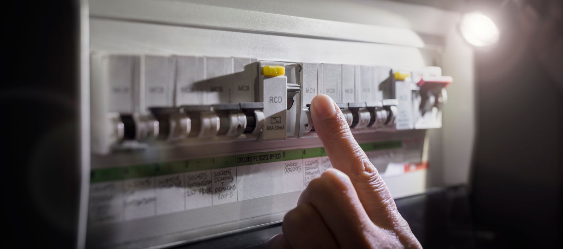Person's hand touching a tripped circuit breaker in a dimly lit electrical panel, illuminated by a flashlight.