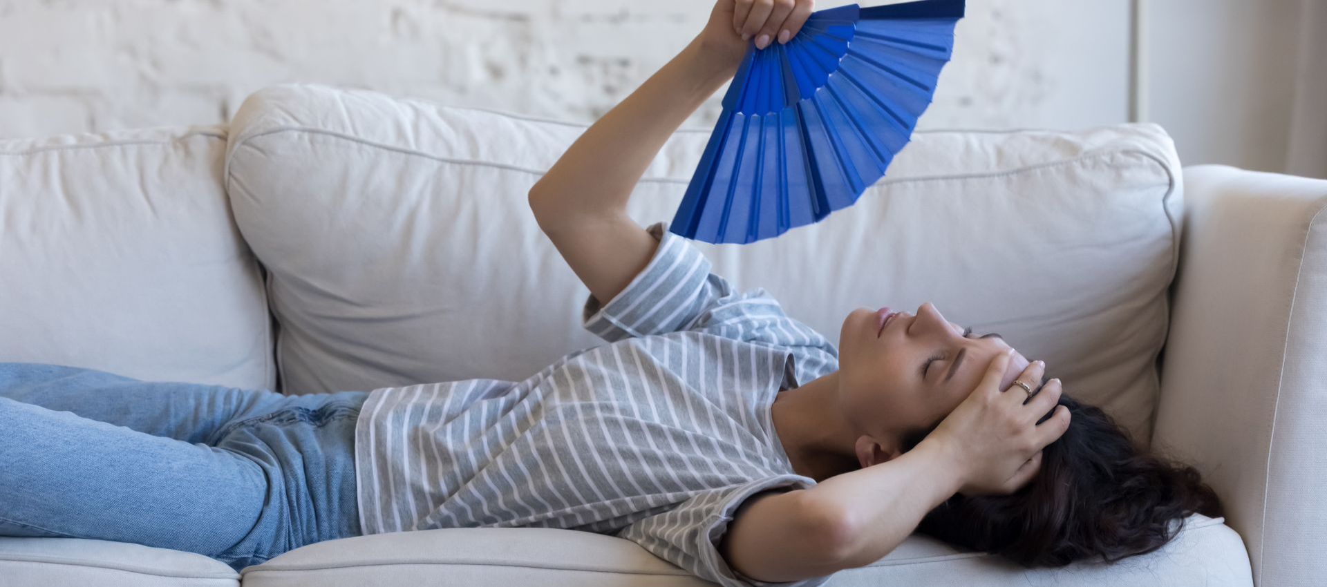 Woman lying on couch, fanning herself with a blue fan, looking hot, indoors.