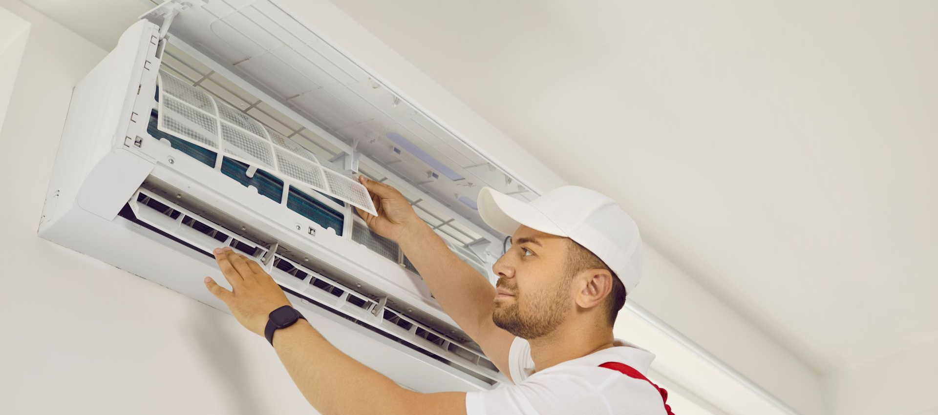 Man in white cap and uniform servicing an air conditioner on a white wall.