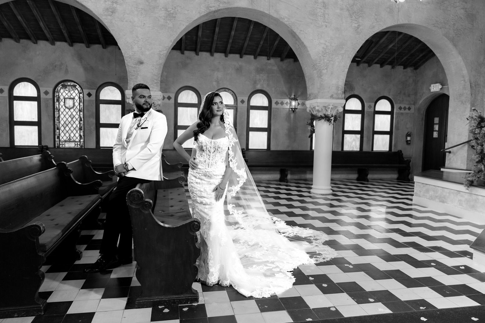 Bride and groom posing in a church-like hall with arched windows and patterned tile floor