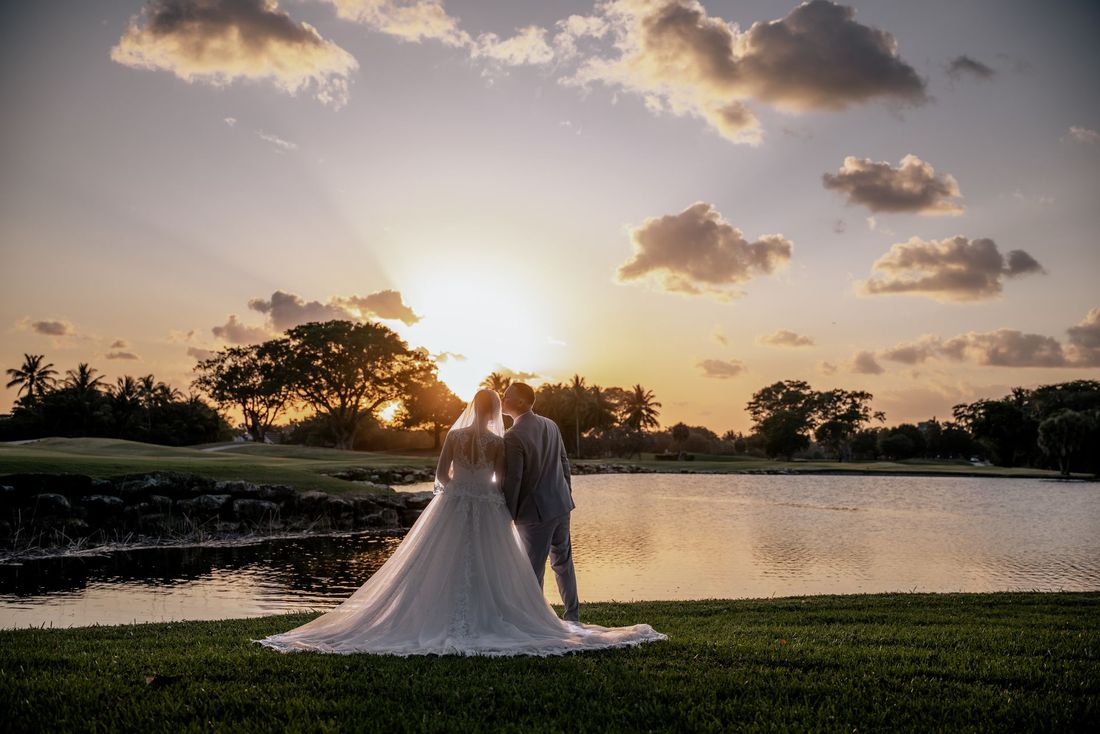 Couple in wedding attire seated on driftwood at sunset beside the ocean, with flowing dress in the breeze