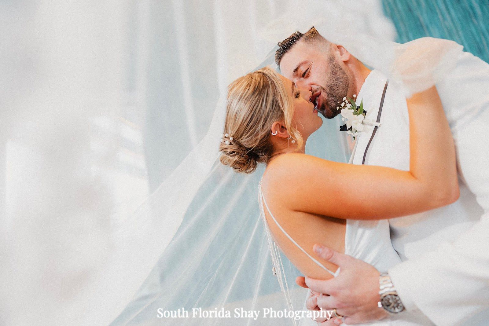 Wedding couple embracing in white attire, foreheads touching by a sheer white curtain