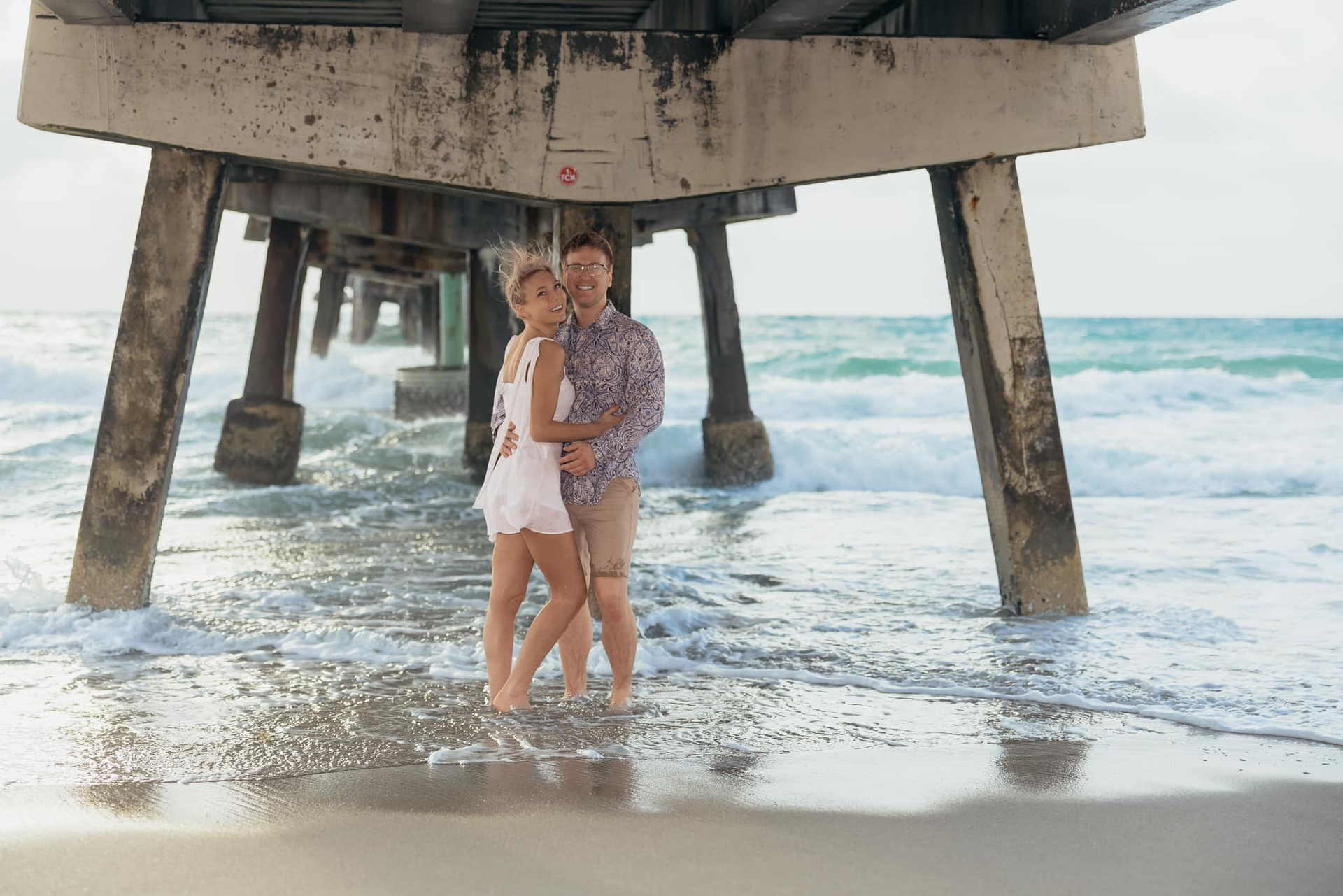 Couple standing under a pier at the shoreline, waves washing around their feet.