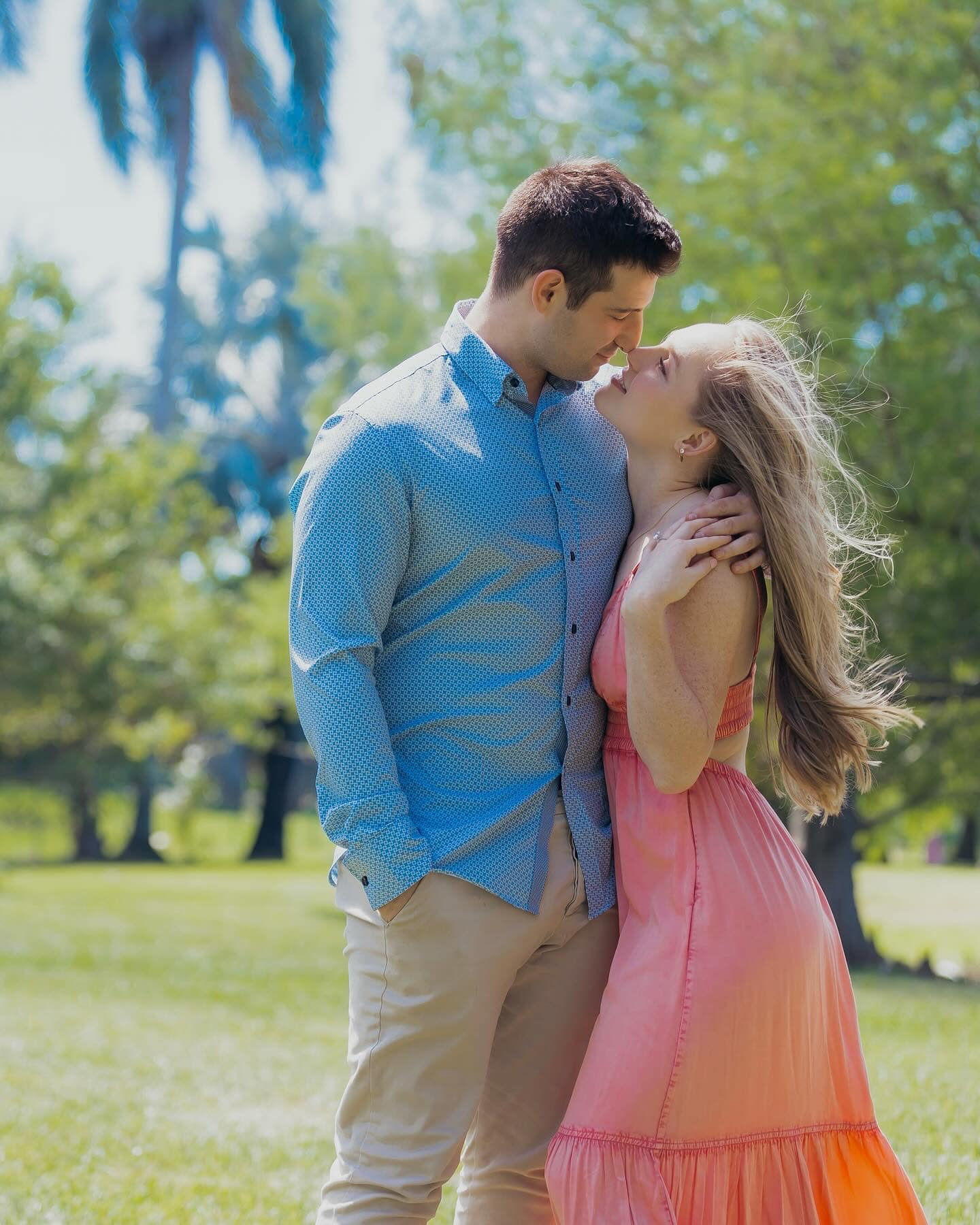 Couple embracing and kissing in a sunny park, the woman in a coral dress and the man in a blue shirt
