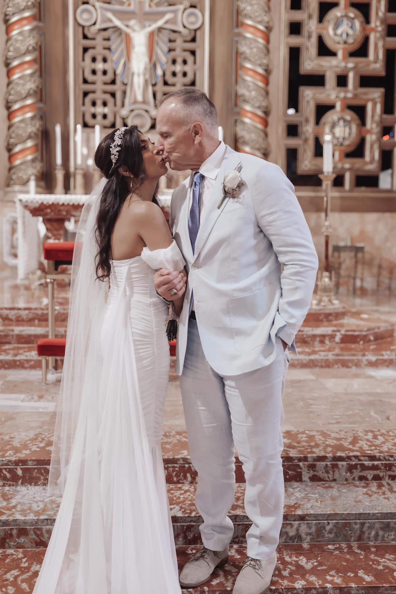 Couple in white wedding attire kissing in an ornate church interior.