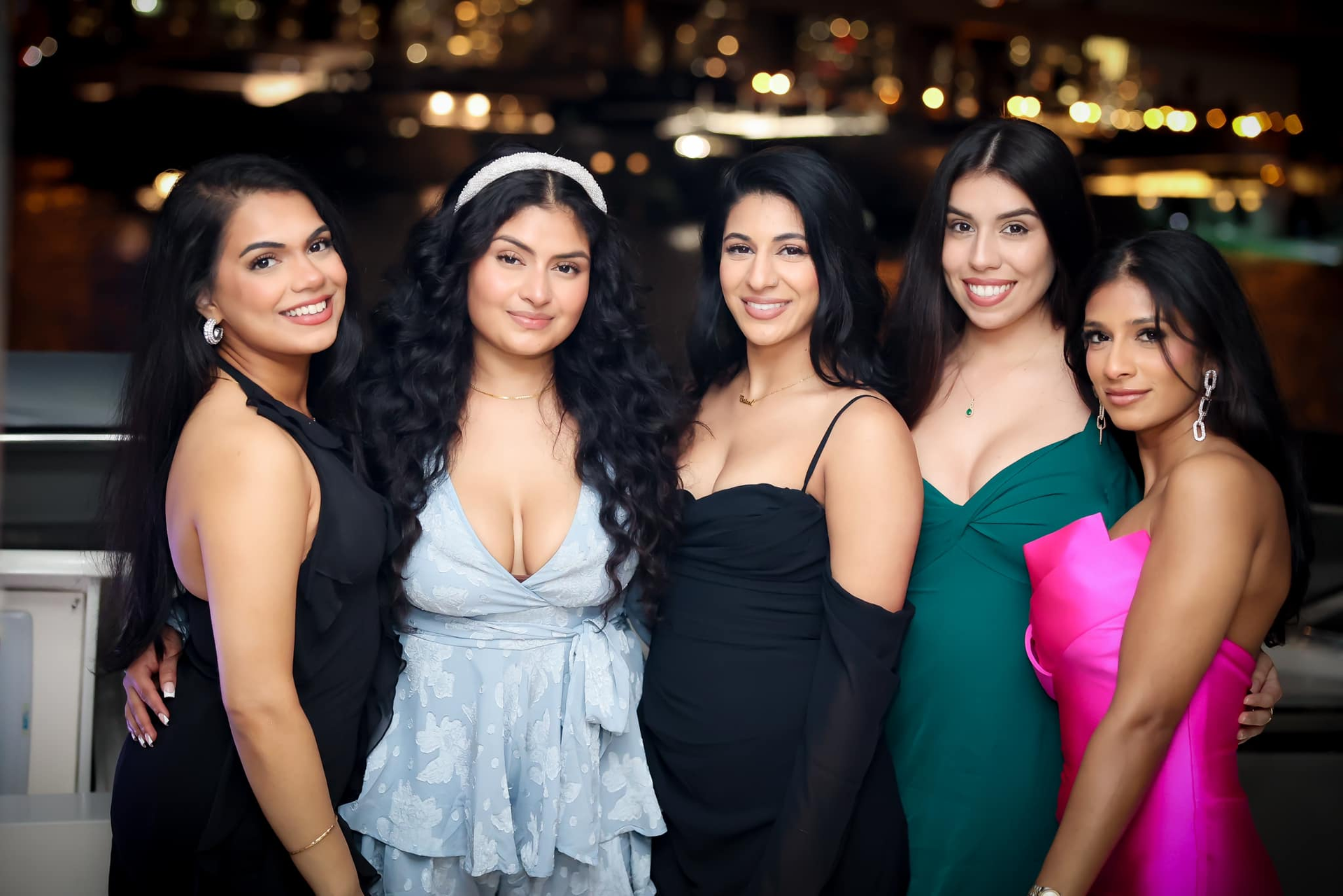 Five women posing together at an evening event, wearing black, light blue, teal, and pink dresses.