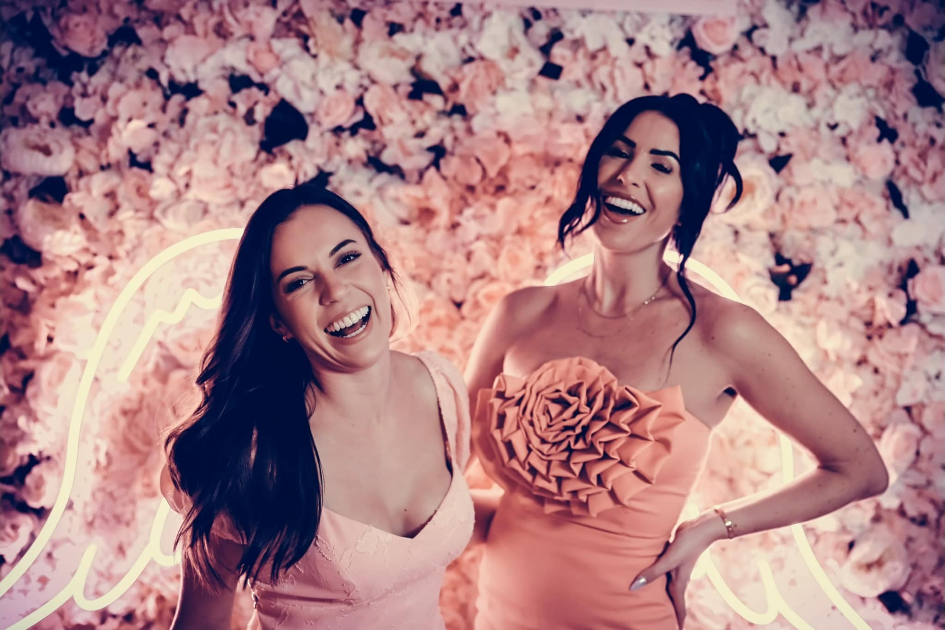 Two smiling women in pink dresses posing before a pink floral wall with neon lights