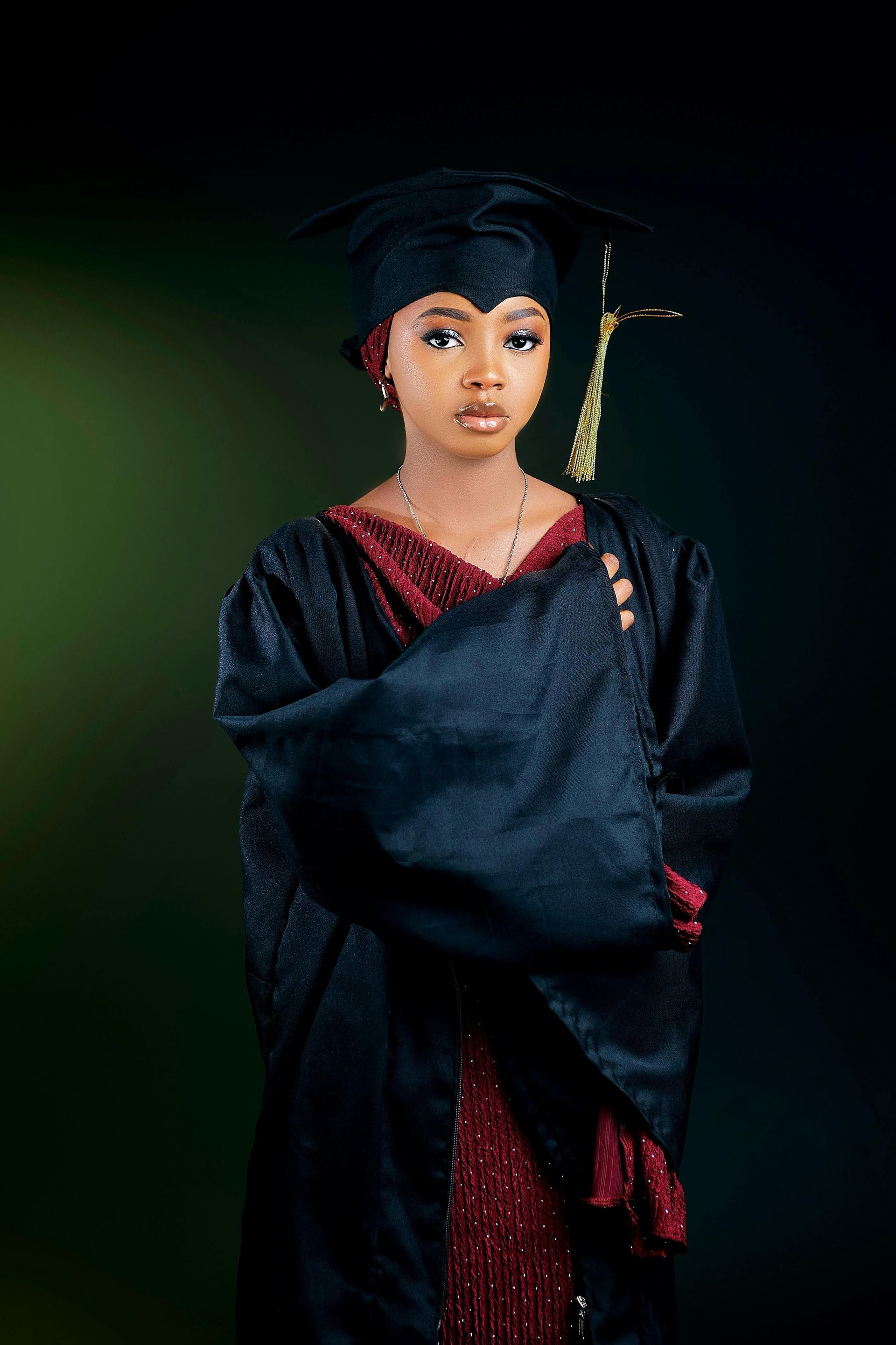 Person in black graduation gown and cap against a dark green background.