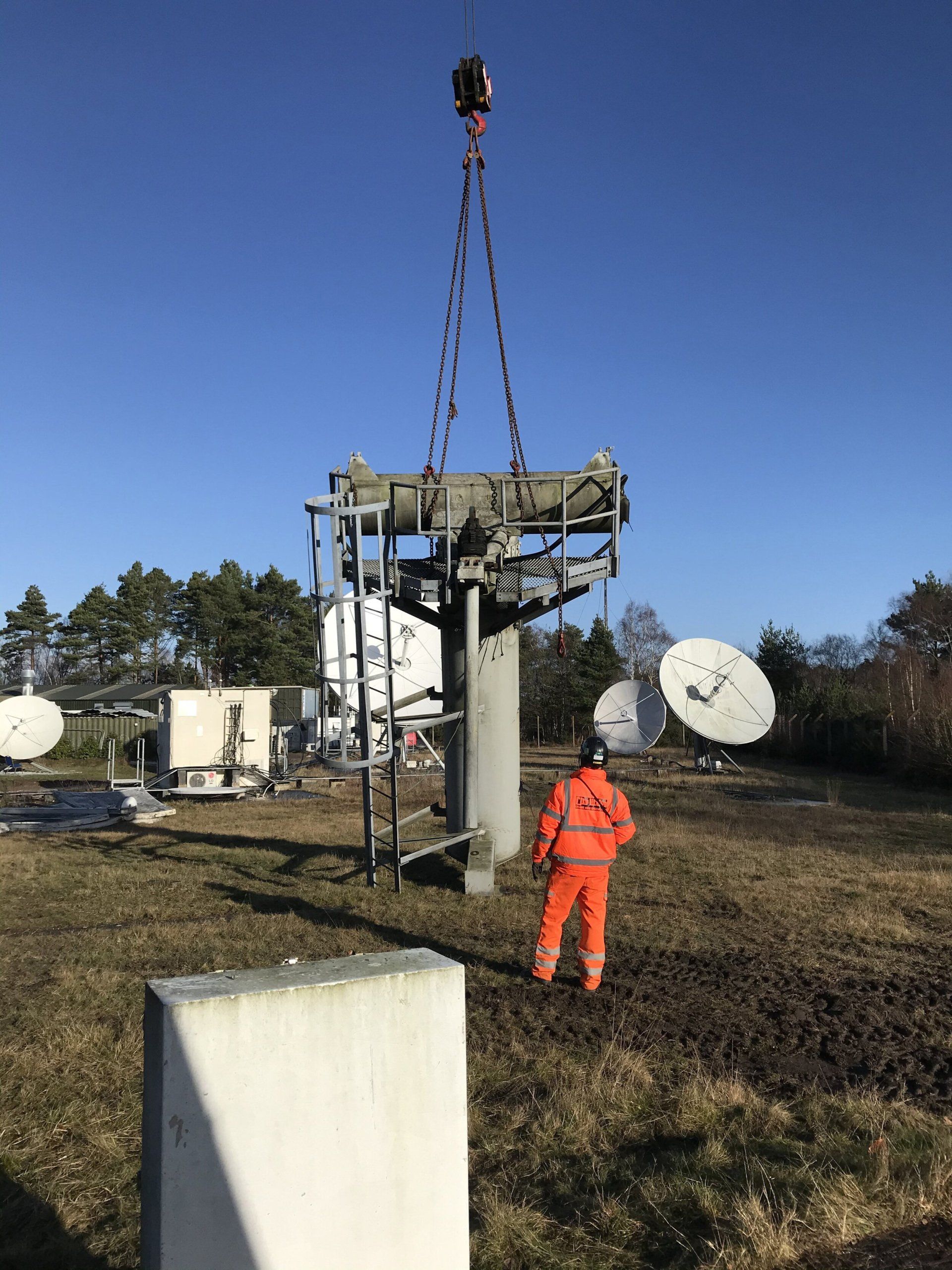 Working in orange over clothes, watching a crane lift an antenna part. 