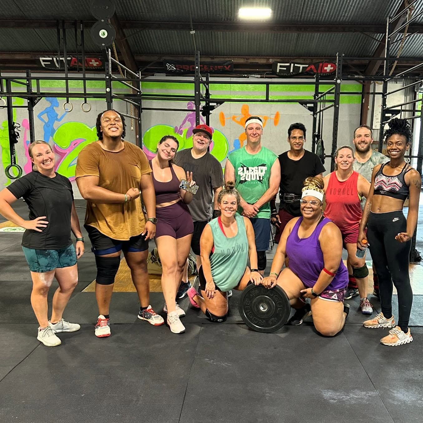 Group workout class posing in a gym with colorful wall art and fitness equipment.