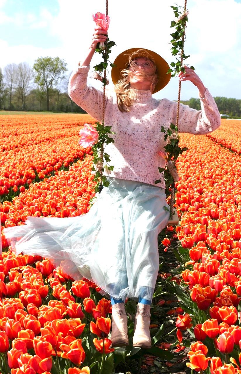 A person in a sun hat, light sweater, and flowy skirt sitting on a flower-decorated swing in a field of orange tulips.