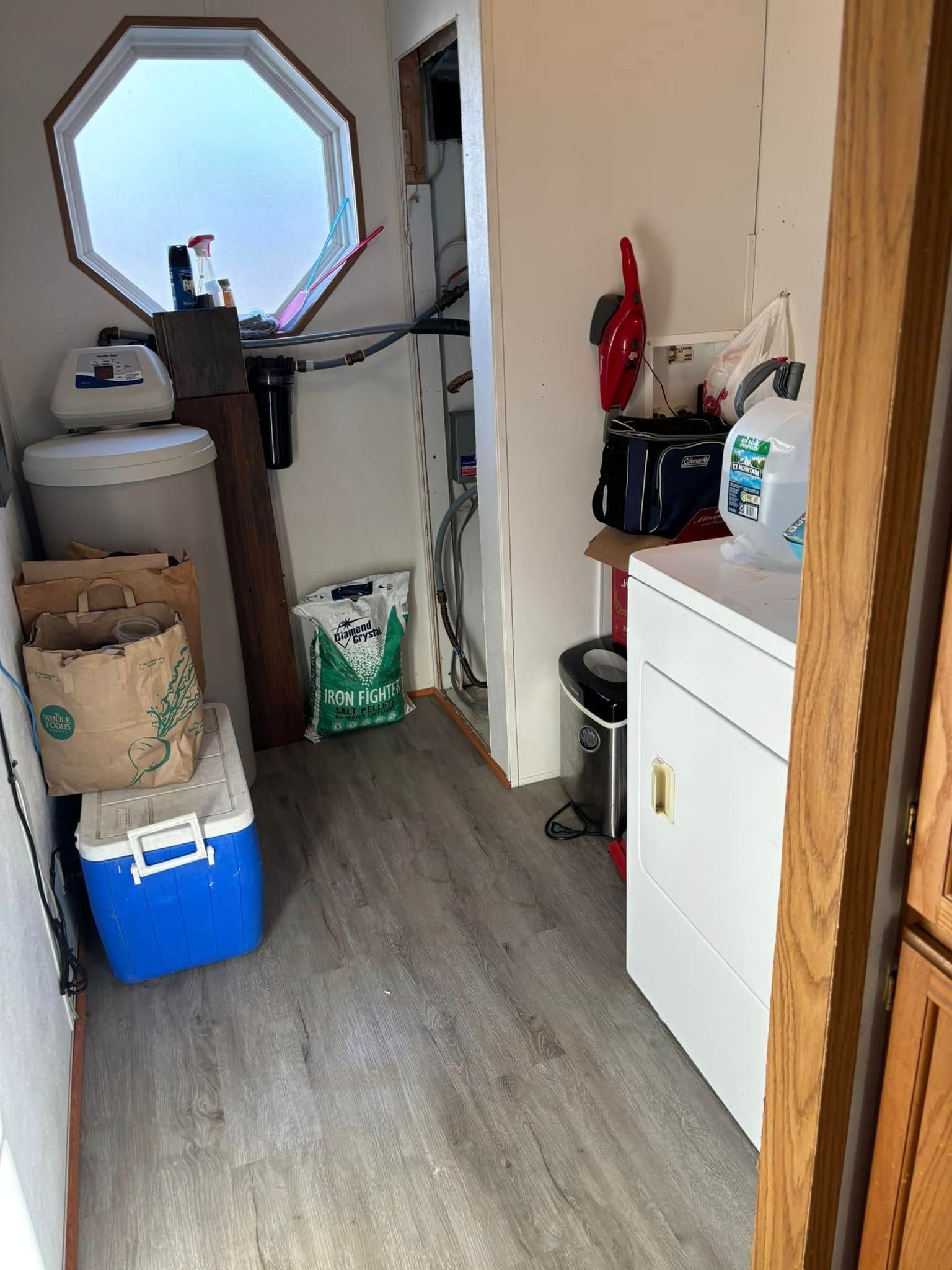 A narrow utility room with a wood-look floor, a water softener, a cooler, a small window, and a white cabinet.