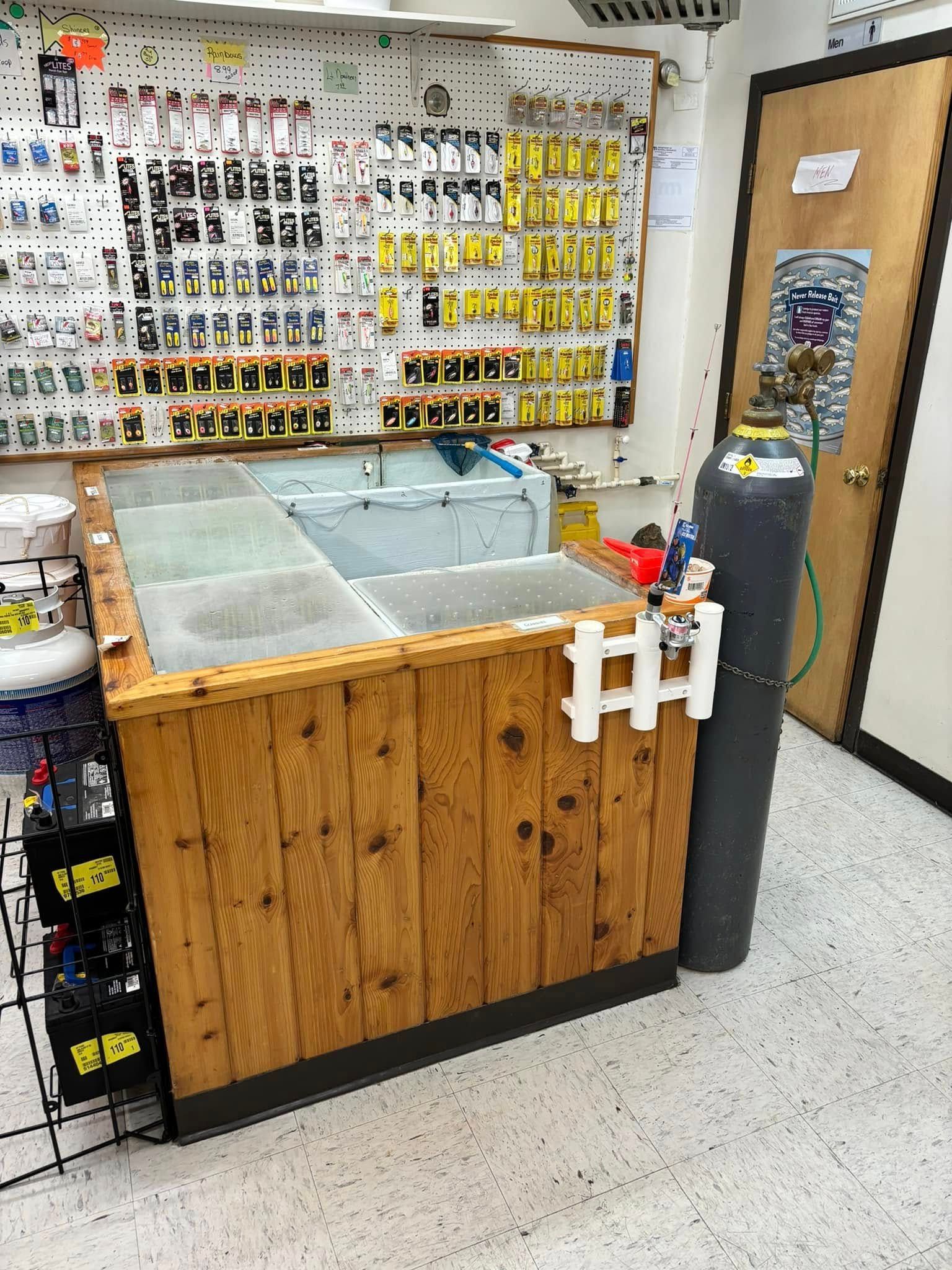 A wooden bait and tackle shop counter with a tall gas cylinder nearby and pegboard display walls filled with supplies.