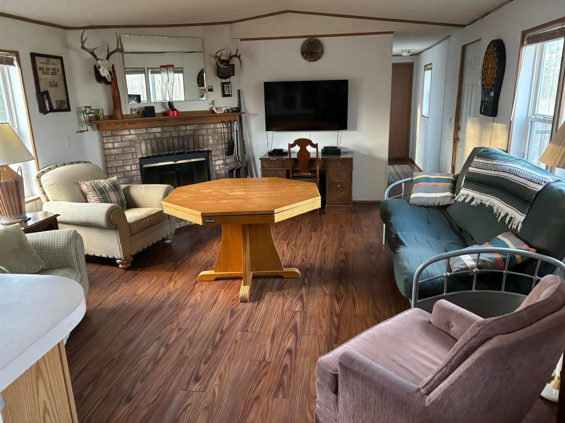 A living room with wood-look flooring, a brick fireplace, an octagonal dining table, a TV, a green couch, and armchairs.