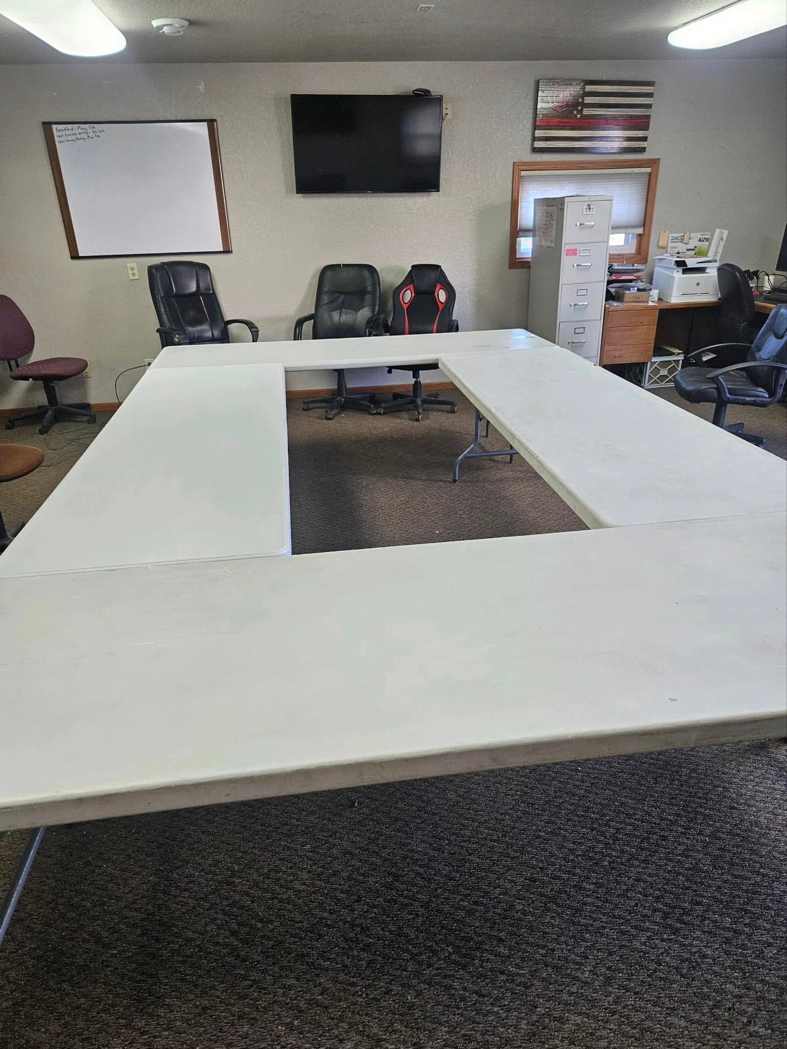 An office meeting room featuring a U-shaped white conference table, several chairs, a whiteboard, and a wall-mounted TV.