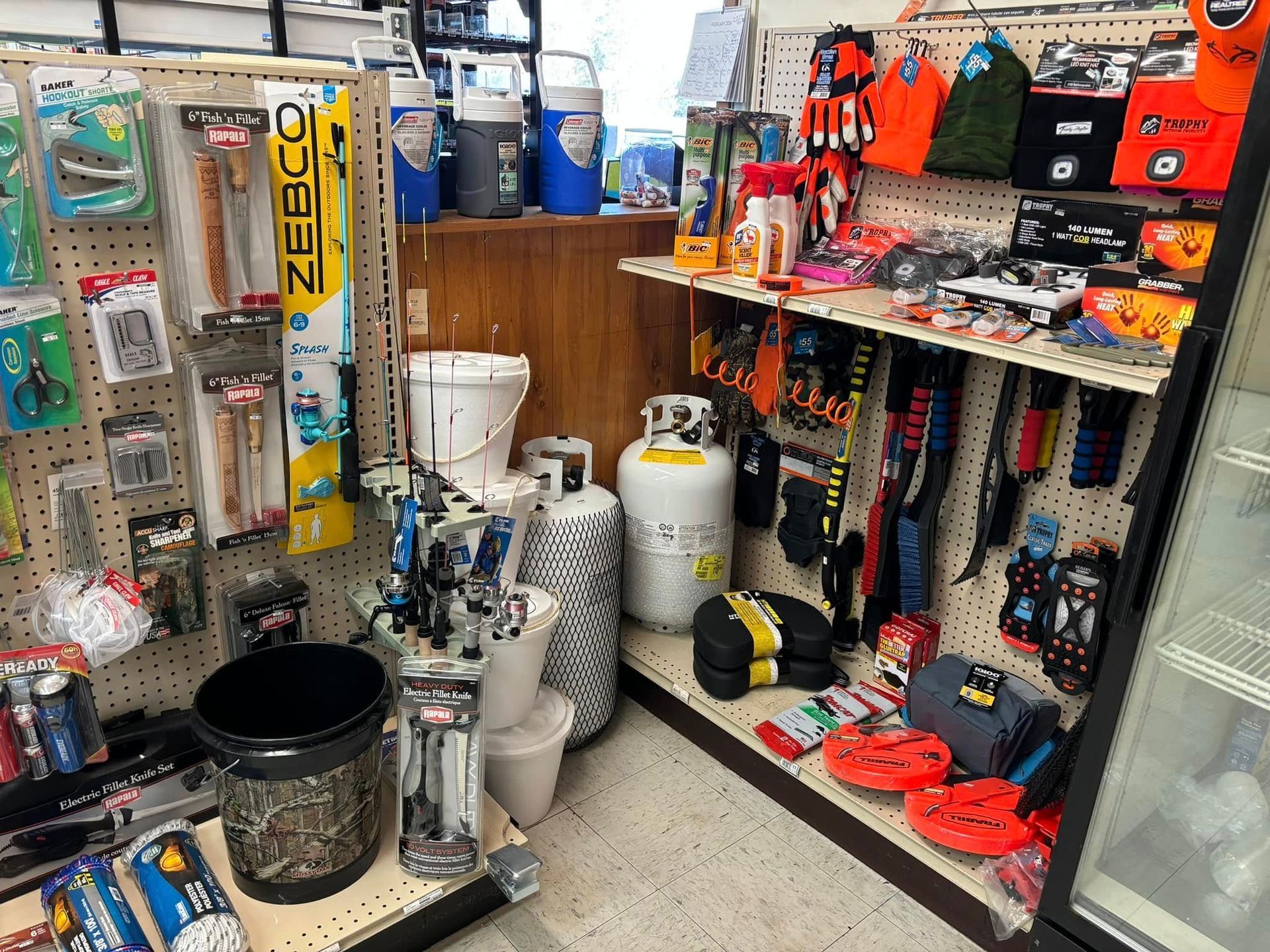A well-stocked aisle in a fishing and outdoors store featuring pegboard displays of gear, propane tanks, and supplies.