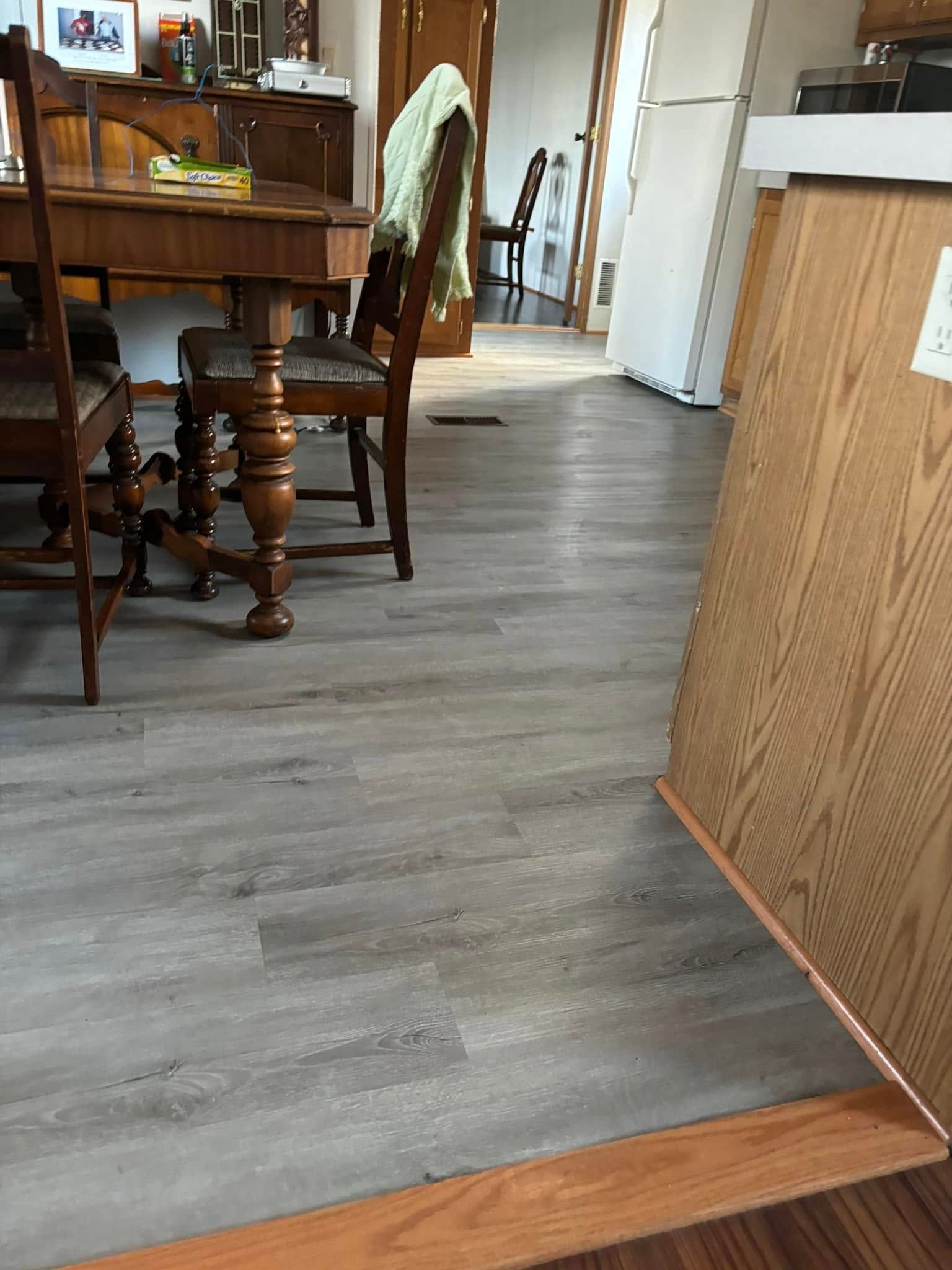 A dining area with wooden furniture on a gray wood-look floor, adjacent to a kitchen counter and a doorway to another room.