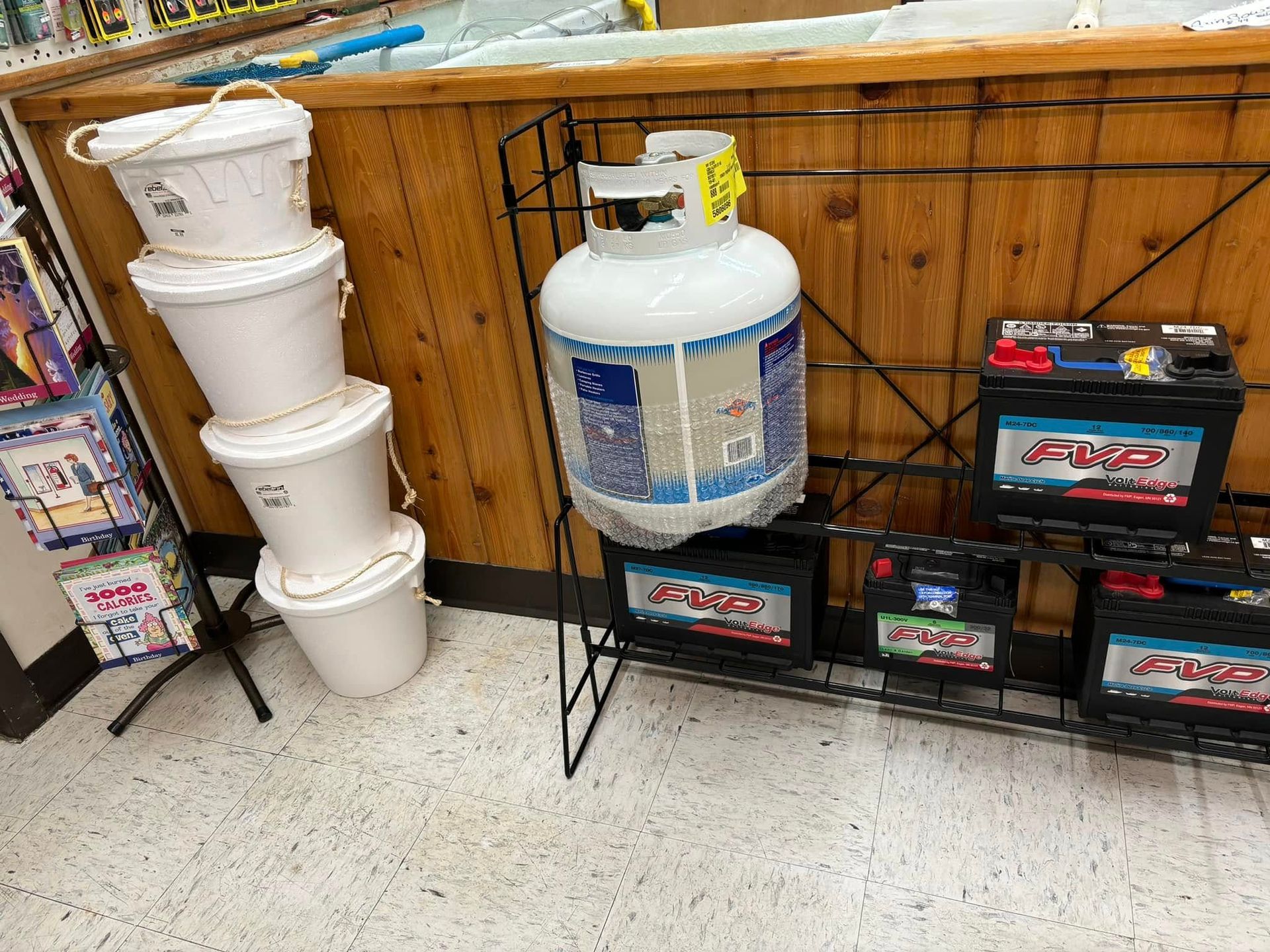 A stack of white plastic buckets stands next to a metal rack displaying a propane tank and several lead-acid batteries.