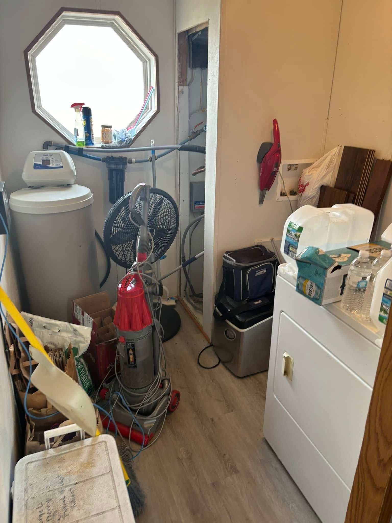A cluttered laundry room with a water softener, vacuum, laundry appliances, and an octagonal window on the back wall.