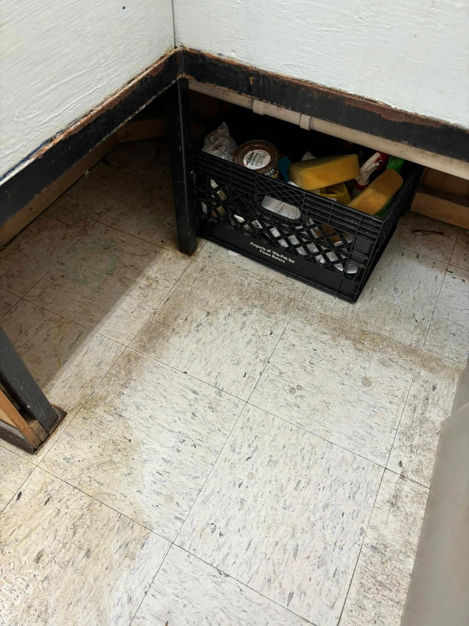 A black plastic crate filled with cleaning supplies sits on a speckled floor under a kitchen or bathroom cabinet frame.