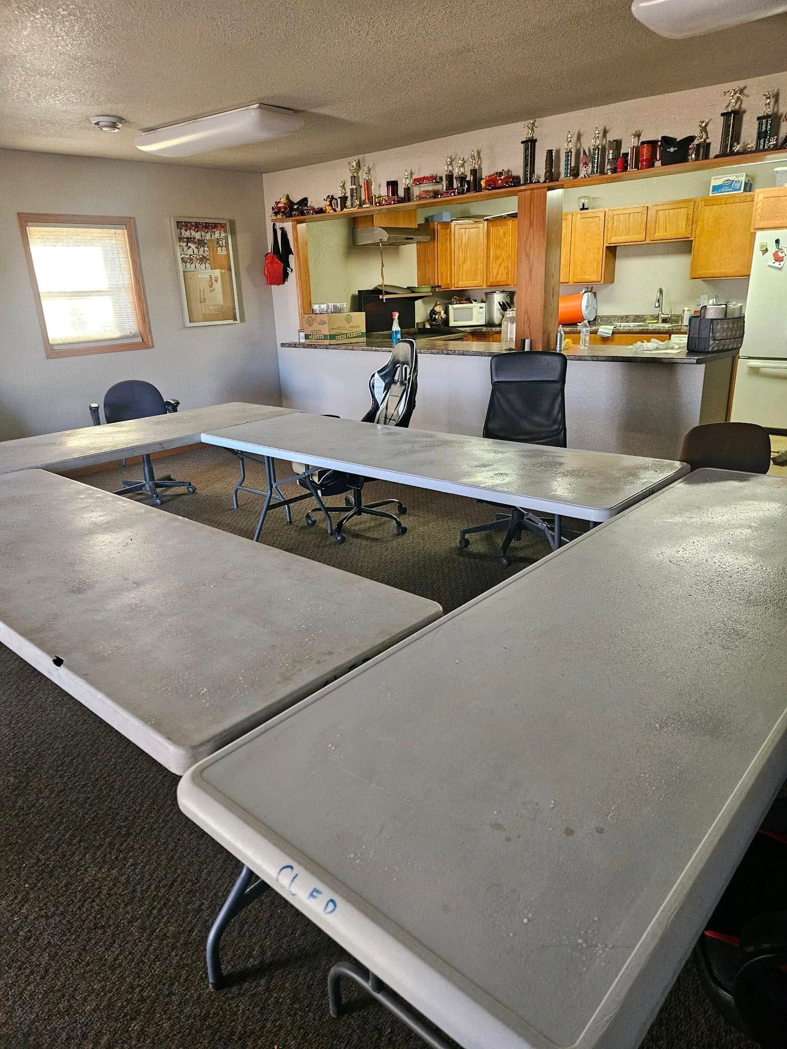A U-shaped configuration of gray folding tables in a meeting room, with chairs, a kitchenette, and shelves of trophies.